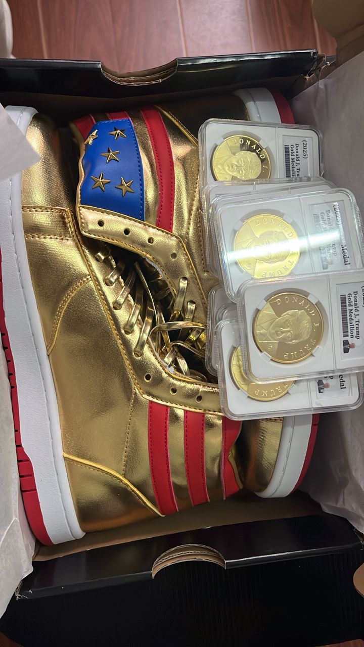 Gold Trump high top sneakers with red stripes and stars beside encased gold Trump coins in box